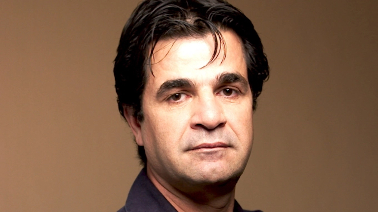 Director Jafar Panahi is pictured looking seriously straight into the camera. The background is brown.
