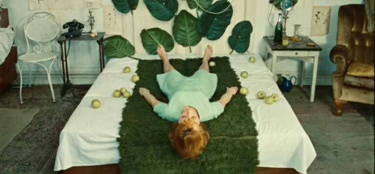 Kuva elokuvasta. Päähenkilö makaa sängyllä, jossa on tekoruohoa ja vihreitä omenoita.