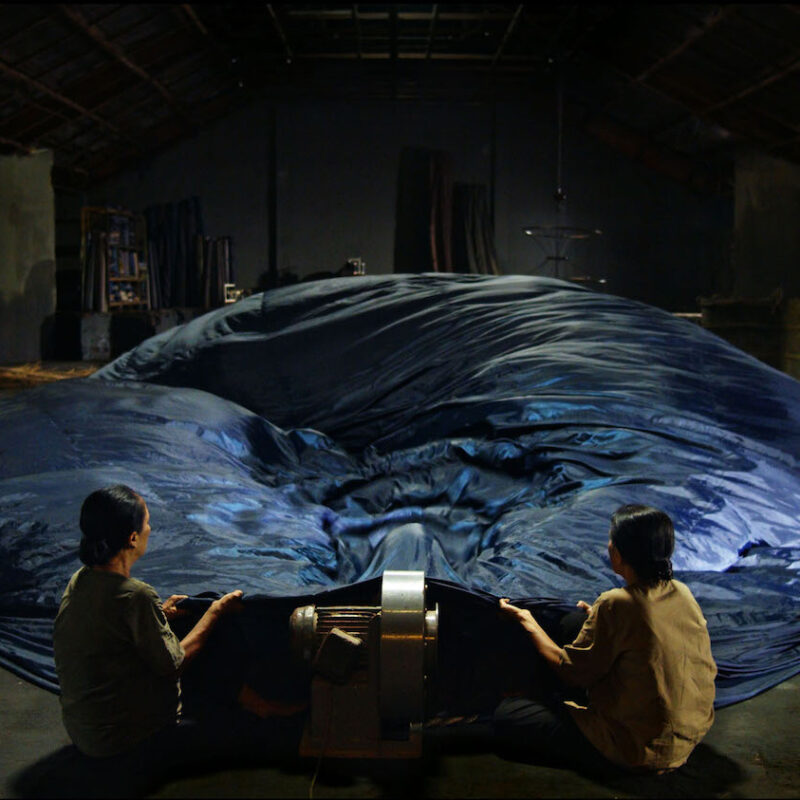 Still-kuva elokuvasta Taste. Kaksi ihmistä heiluttaa suurta sinistä kangasta.